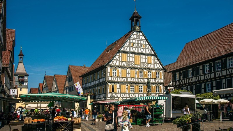 Der Marktplatz in Winnenden zeigt Fachwerkhäuser und Marktstände unter einem klaren blauen Himmel. Menschen schlendern und kaufen ein., © Stuttgart-Marketing GmbH, Sarah Schmid Der Marktplatz in Winnenden zeigt Fachwerkhäuser und Marktstände unter einem klaren blauen Himmel. Menschen schlendern und kaufen ein., © Stuttgart-Marketing GmbH, Sarah Schmid