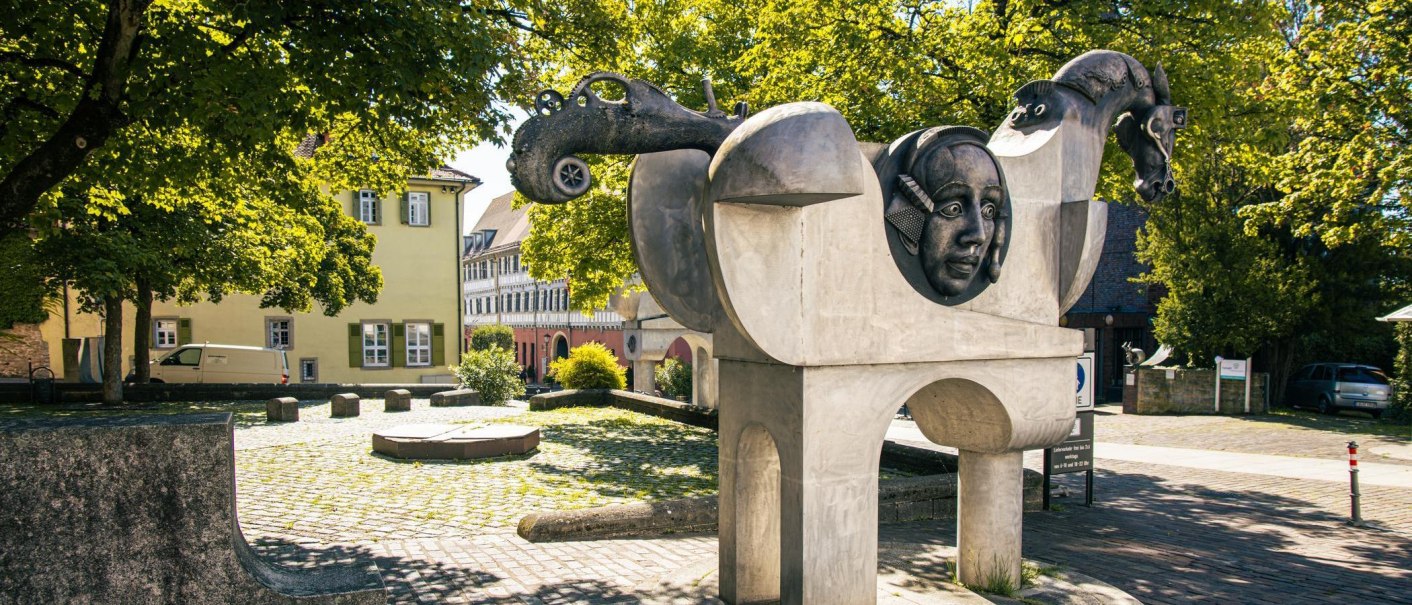 Moderne Skulptur in Bietigheim-Bissingen mit Pferdeköpfen und einem menschlichen Gesicht, umgeben von Bäumen und historischen Gebäuden., © Stuttgart-Marketing GmbH, Sarah Schmid