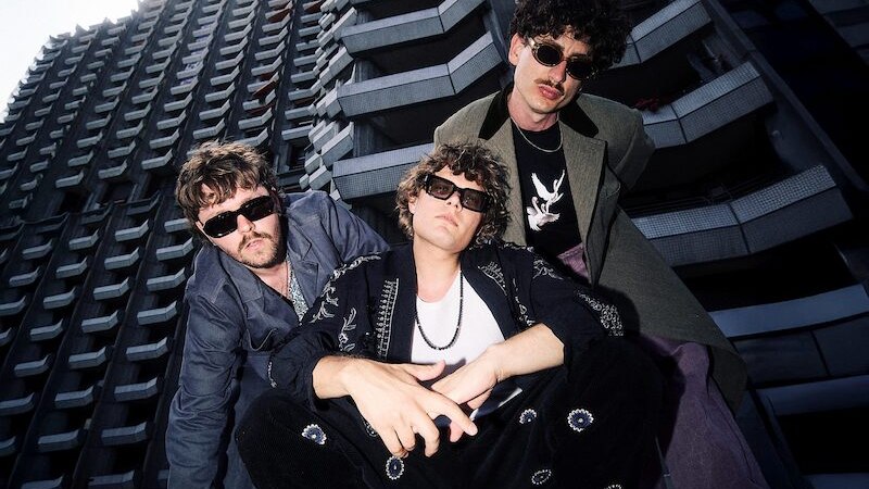 Drei Männer mit Sonnenbrillen posieren lässig vor einem modernen, hohen Gebäude. Sie tragen modische Kleidung und schauen in die Kamera., © Kulturhaus Schwanen Drei Männer mit Sonnenbrillen posieren lässig vor einem modernen, hohen Gebäude. Sie tragen modische Kleidung und schauen in die Kamera., © Kulturhaus Schwanen