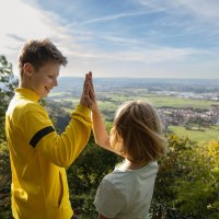 Zwei Kinder geben sich ein High-Five auf einem Hügel mit Blick auf eine weite Landschaft und einen blauen Himmel., © Foto Thomas Zehnder