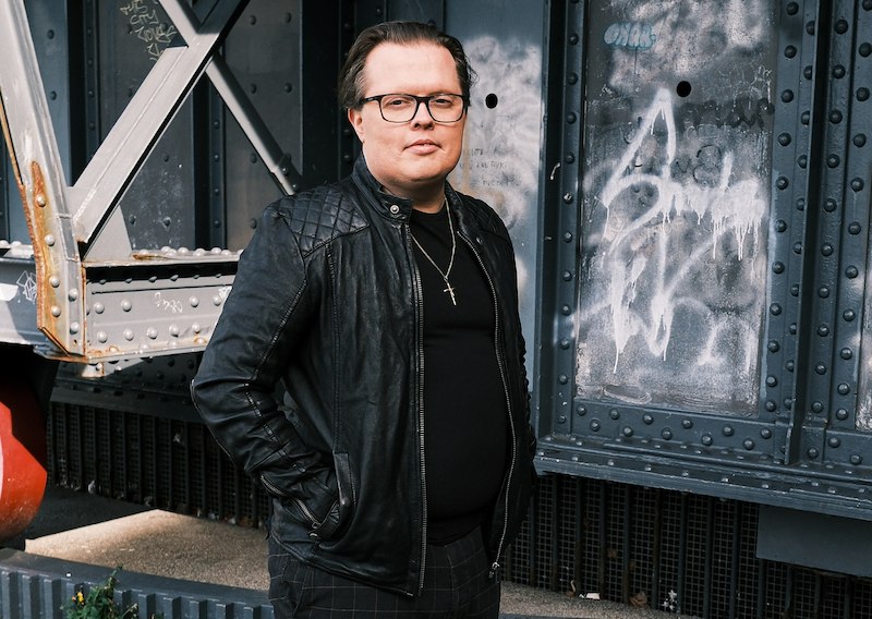 Person in schwarzer Lederjacke und Brille steht vor einer metallischen Wand mit Graffiti., © Scala Kultur Live gGmbH