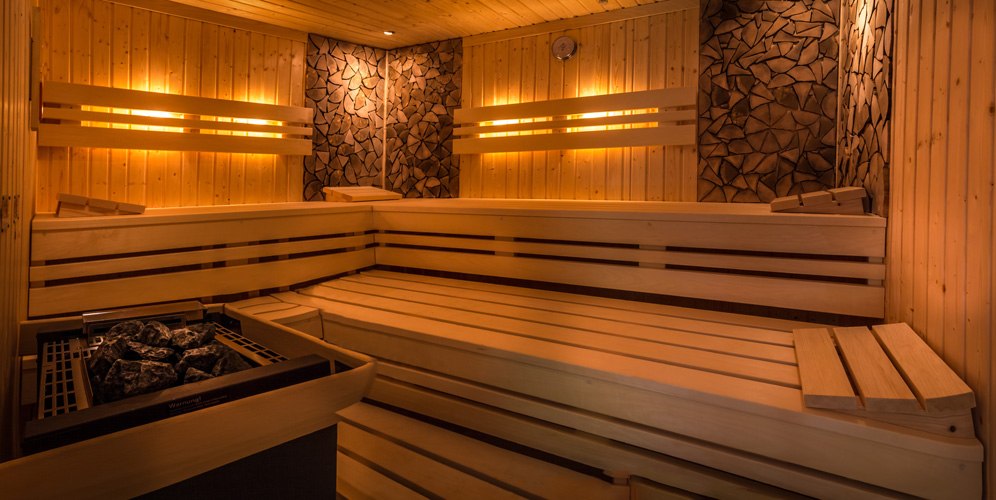 Moderne Sauna mit Holzbänken, warmem Licht und einem Ofen mit Steinen. Die Wände sind aus Holz und dekorativem Stein gestaltet., © Kulinarium an der Glems