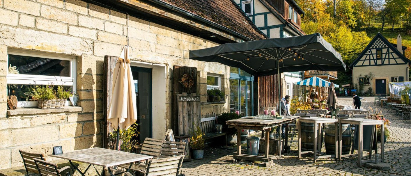 Gemütlicher Café-Außenbereich mit Holzmöbeln und Sonnenschirm, umgeben von Fachwerkhäusern und herbstlicher Landschaft., © Stuttgart-Marketing GmbH, Sarah Schmid