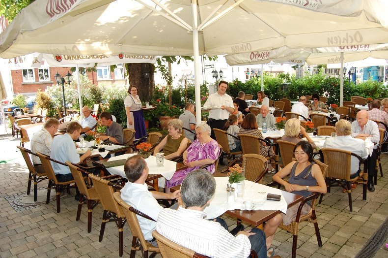 Ein belebter Biergarten mit vielen Menschen, die an Tischen unter großen Sonnenschirmen sitzen. Kellner bedienen die Gäste. Es ist ein sonniger Tag., © Jeanette Bender Ein belebter Biergarten mit vielen Menschen, die an Tischen unter großen Sonnenschirmen sitzen. Kellner bedienen die Gäste. Es ist ein sonniger Tag., © Jeanette Bender