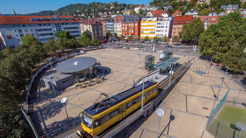 Luftaufnahme des Marienplatzes in Stuttgart. Eine gelbe Straßenbahn fährt vorbei, umgeben von Bäumen und Gebäuden. Der Platz ist belebt und sonnig., © Stuttgart-Marketing GmbH, Achim Mende Luftaufnahme des Marienplatzes in Stuttgart. Eine gelbe Straßenbahn fährt vorbei, umgeben von Bäumen und Gebäuden. Der Platz ist belebt und sonnig., © Stuttgart-Marketing GmbH, Achim Mende