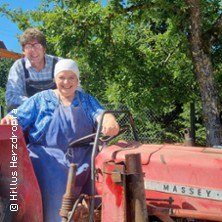 Zwei Personen sitzen auf einem roten Traktor. Im Hintergrund sind grüne Bäume zu sehen. Die Szene wirkt ländlich und idyllisch., © links im Bild