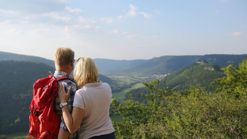 Ein Paar mit rotem Rucksack genießt den Ausblick vom Eppenzillfelsen auf eine grüne Hügellandschaft unter blauem Himmel., © Bad urach Tourismus