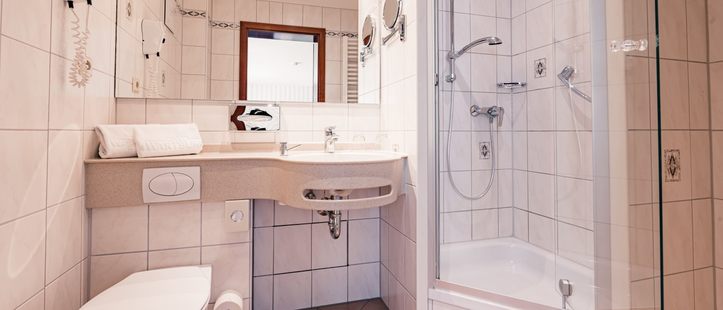 Modernes Badezimmer mit Dusche, Waschbecken und Toilette. Weiße Fliesen, Spiegel und Handtücher sind sichtbar. Ein Föhn hängt an der Wand., © TOMAS