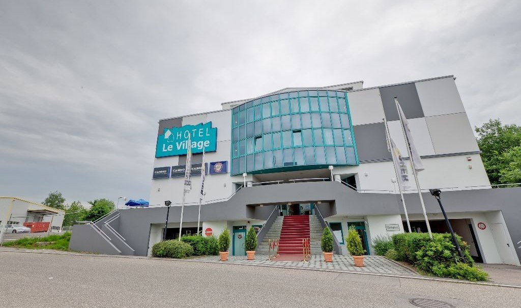 Das Hotel Le Village zeigt eine moderne Fassade mit Glasfront und Treppe. Vor dem Gebäude wehen Fahnen, der Himmel ist bewölkt., © Hotel Le Village Das Hotel Le Village zeigt eine moderne Fassade mit Glasfront und Treppe. Vor dem Gebäude wehen Fahnen, der Himmel ist bewölkt., © Hotel Le Village