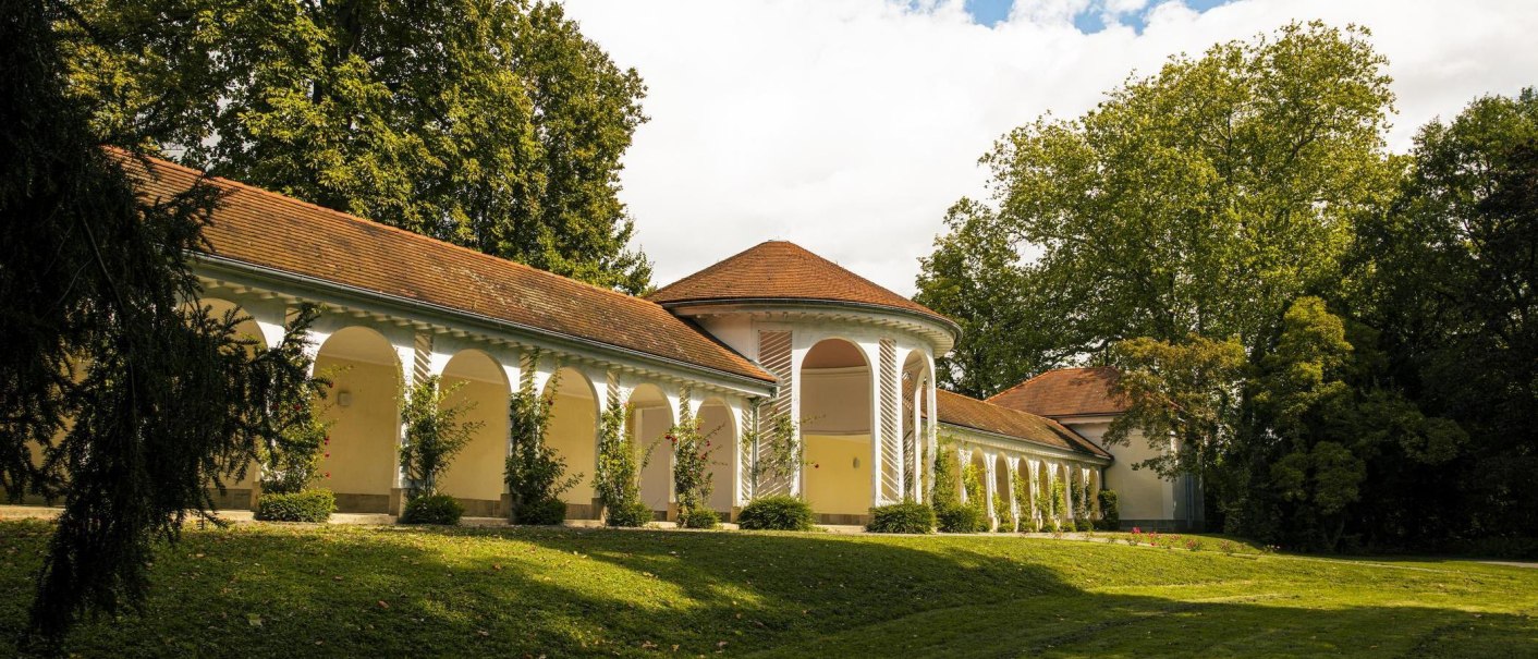 Die Wandelhalle in Bad Boll zeigt elegante Arkaden und ein rotes Ziegeldach, eingebettet in eine gr&uuml;ne Landschaft mit B&auml;umen und Rasen., &copy; SMG, Sarah Schmid
