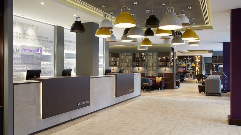 Moderne Hotel-Lobby mit Rezeption, hängenden Lampen und gemütlichem Sitzbereich. An der Wand steht "Premier Inn". Im Hintergrund ist eine Lounge sichtbar., © Premier Inn