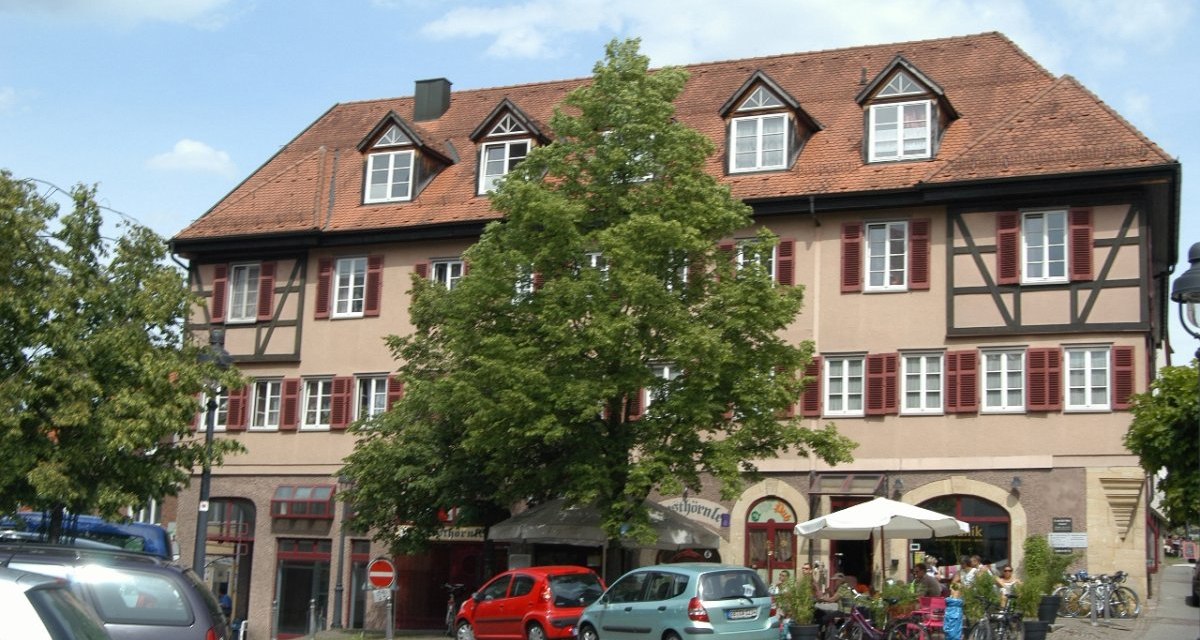 Fachwerkhaus mit roten Fensterl&auml;den, geparkte Autos und Fahrr&auml;der. Menschen sitzen unter Sonnenschirmen vor einem Caf&eacute;. B&auml;ume im Vordergrund., &copy; Natur.Nah. Sch&ouml;nbuch & Heckeng&auml;u