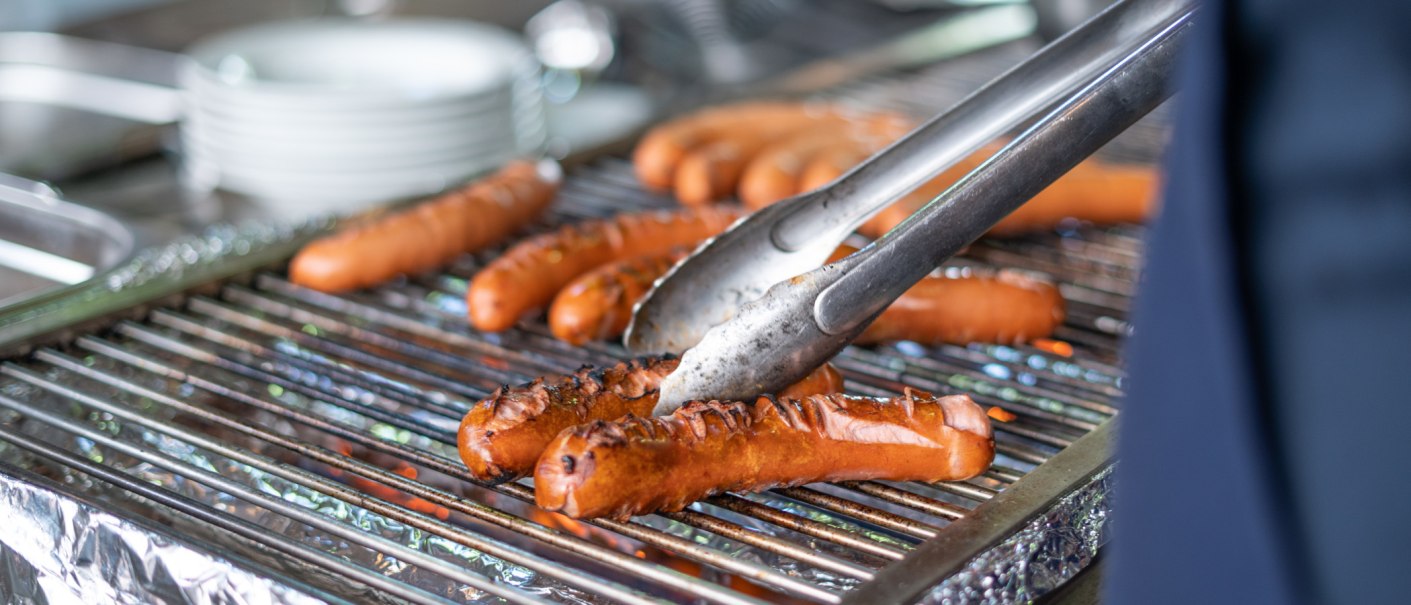 Würstchen werden auf einem Grill mit einer Zange gewendet. Im Hintergrund sind unscharfe Teller zu sehen., © Frank Nonnenmann Würstchen werden auf einem Grill mit einer Zange gewendet. Im Hintergrund sind unscharfe Teller zu sehen., © Frank Nonnenmann