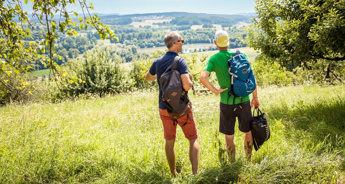 Zwei Männer mit Rucksäcken stehen auf einer Wiese und schauen in die grüne Landschaft unter blauem Himmel., © hochgehberge Zwei Männer mit Rucksäcken stehen auf einer Wiese und schauen in die grüne Landschaft unter blauem Himmel., © hochgehberge