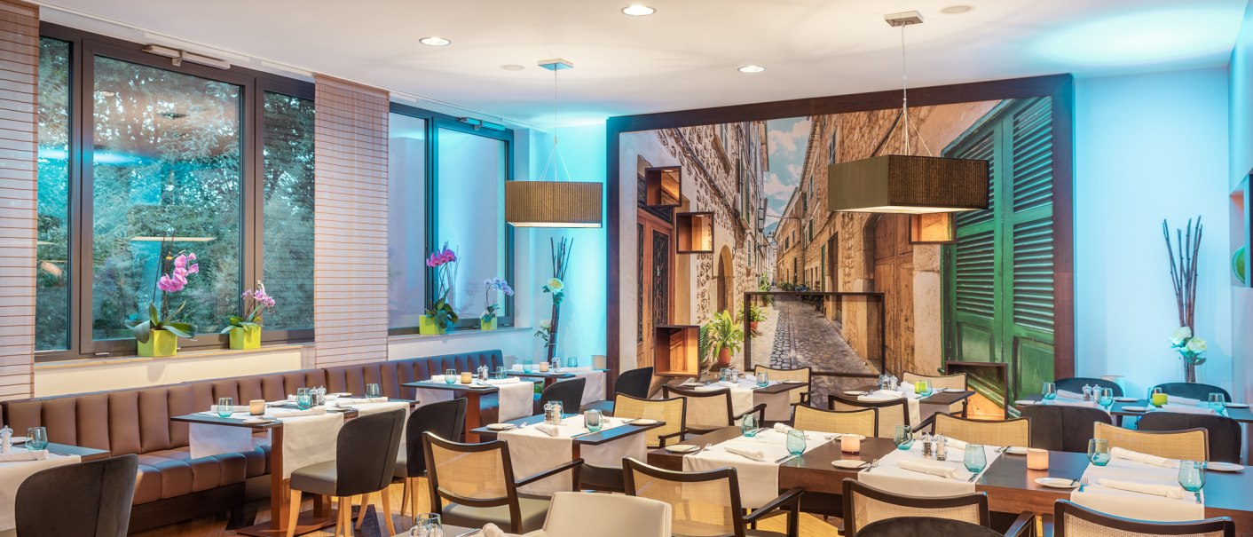 Elegantes Restaurant mit modernen Tischen, Stühlen und Wandbild einer Gasse. Dezente Beleuchtung und dekorative Pflanzen schaffen eine gemütliche Atmosphäre., © NH Hotel Stuttgart Airport