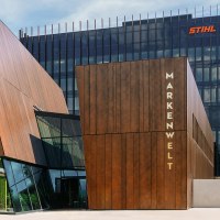 Moderne Gebäude der STIHL Markenwelt mit Holzverkleidung und Glasfronten, umgeben von Bäumen. Sonnenschirme und Sitzgelegenheiten im Vordergrund., © STIHL Markenwelt