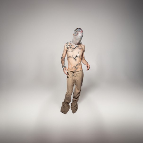 Person mit Maske und Pflanzen auf dem Oberkörper, trägt eine beige Hose in einem neutralen Raum., © Scala Kultur Live gGmbH