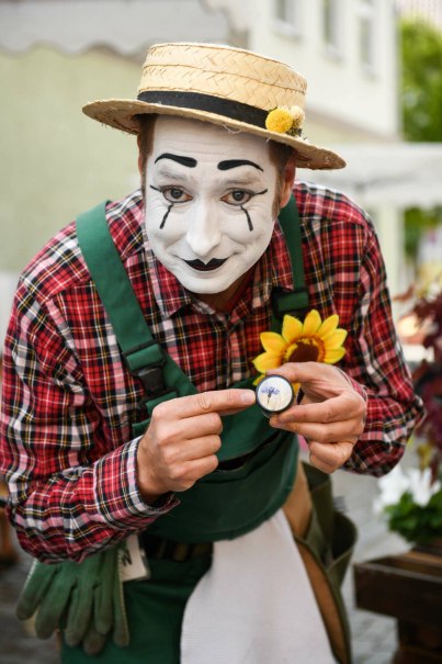 Ein Pantomime mit weißem Gesicht und Strohhut zeigt auf eine Lupe mit Blume. Er trägt ein kariertes Hemd und grüne Hosenträger.