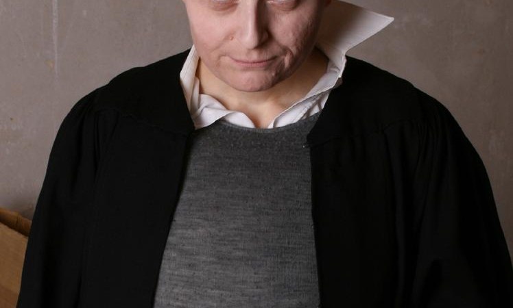 Eine Person mit Glatze und Kunstblut auf der Stirn hält einen Krug mit der Aufschrift 'Gruß aus...'. Sie trägt einen schwarzen Umhang und ein graues Oberteil., © Foto_(c)_Dirk Holtkamp Endemann Eine Person mit Glatze und Kunstblut auf der Stirn hält einen Krug mit der Aufschrift 'Gruß aus...'. Sie trägt einen schwarzen Umhang und ein graues Oberteil., © Foto_(c)_Dirk Holtkamp Endemann