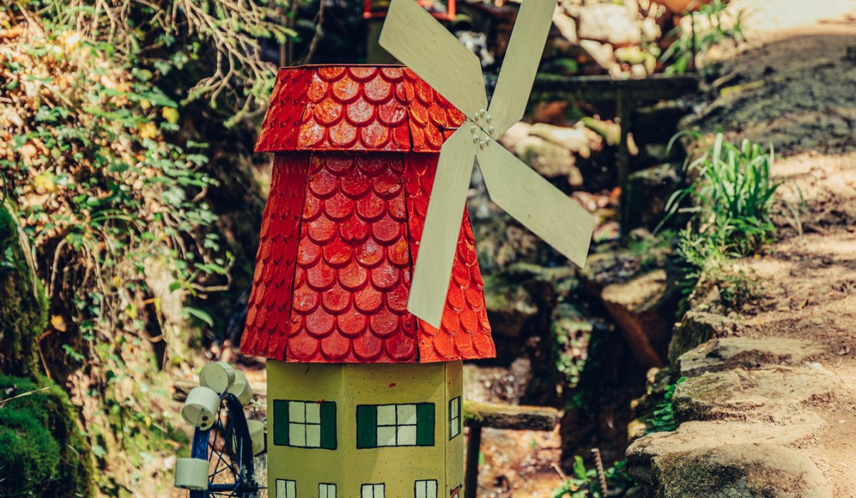 Eine kleine, bunte Mühle mit rotem Dach und Wasserrad steht im Wald neben einem Pfad. Im Hintergrund sind Bäume und Spaziergänger zu sehen., © Tourismus Ostalb