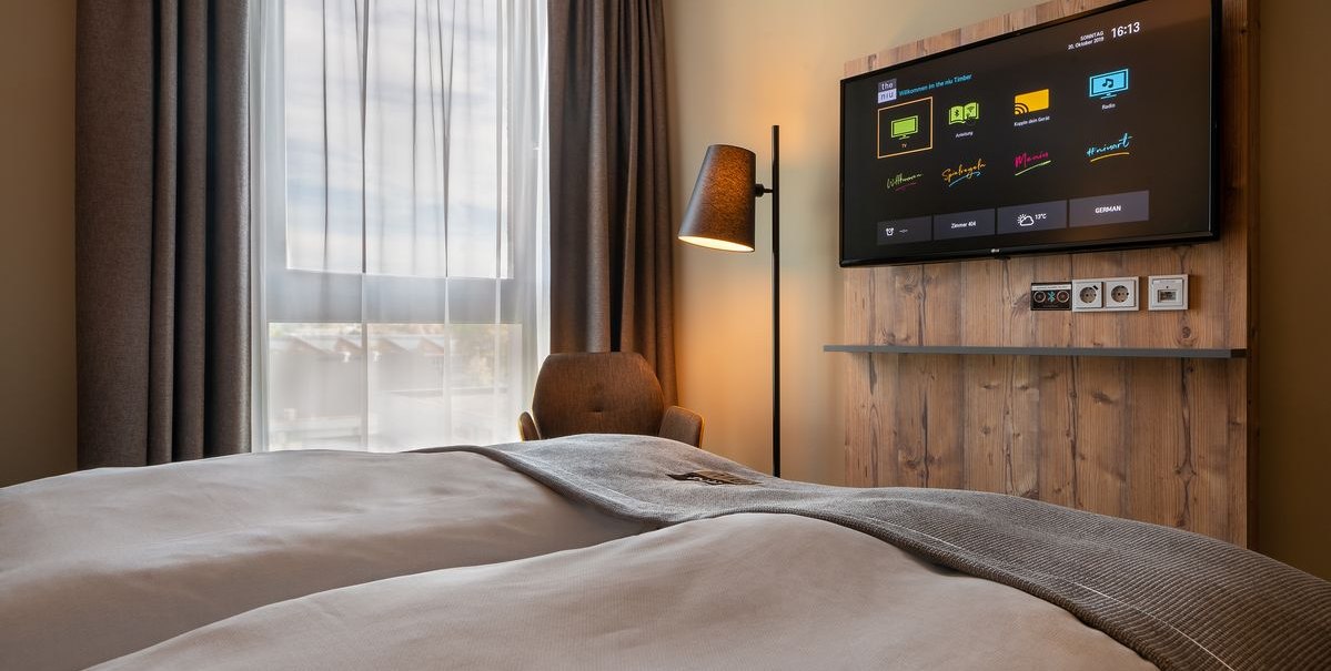 Modernes Hotelzimmer mit Doppelbett, großem Fenster, Fernseher an Holzwand und Stehlampe., © Novum Hospitality Modernes Hotelzimmer mit Doppelbett, großem Fenster, Fernseher an Holzwand und Stehlampe., © Novum Hospitality