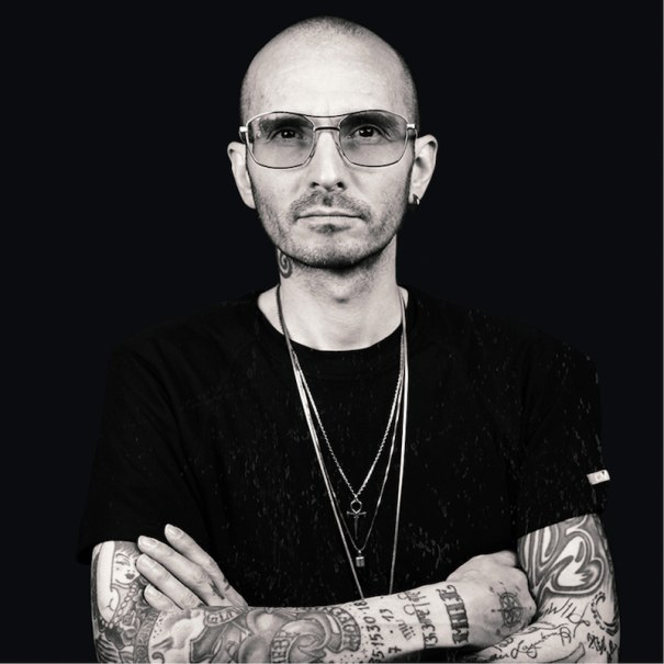 Person mit Glatze, Sonnenbrille und tätowierten Armen vor schwarzem Hintergrund. Sie trägt ein schwarzes T-Shirt und mehrere Halsketten., © Expedition Erde Person mit Glatze, Sonnenbrille und tätowierten Armen vor schwarzem Hintergrund. Sie trägt ein schwarzes T-Shirt und mehrere Halsketten., © Expedition Erde