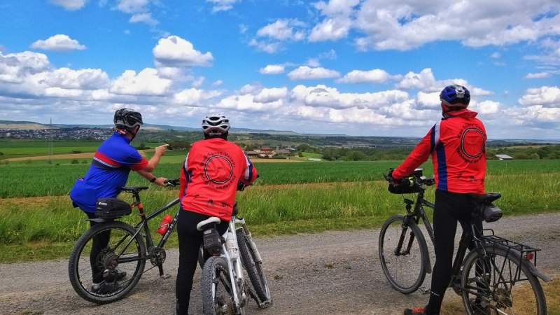 Drei Radfahrer in bunter Sportkleidung stehen auf einem Feldweg und schauen in die weite, grüne Landschaft unter einem blauen Himmel mit Wolken., © Land der 1000 Hügel - Kraichgau-Stromberg Drei Radfahrer in bunter Sportkleidung stehen auf einem Feldweg und schauen in die weite, grüne Landschaft unter einem blauen Himmel mit Wolken., © Land der 1000 Hügel - Kraichgau-Stromberg