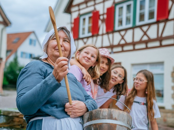 Eine Frau in historischer Kleidung hält einen Holzlöffel, umgeben von vier Kindern. Im Hintergrund ein Fachwerkhaus mit roten Fensterläden., © Stadt Waldenbuch Eine Frau in historischer Kleidung hält einen Holzlöffel, umgeben von vier Kindern. Im Hintergrund ein Fachwerkhaus mit roten Fensterläden., © Stadt Waldenbuch