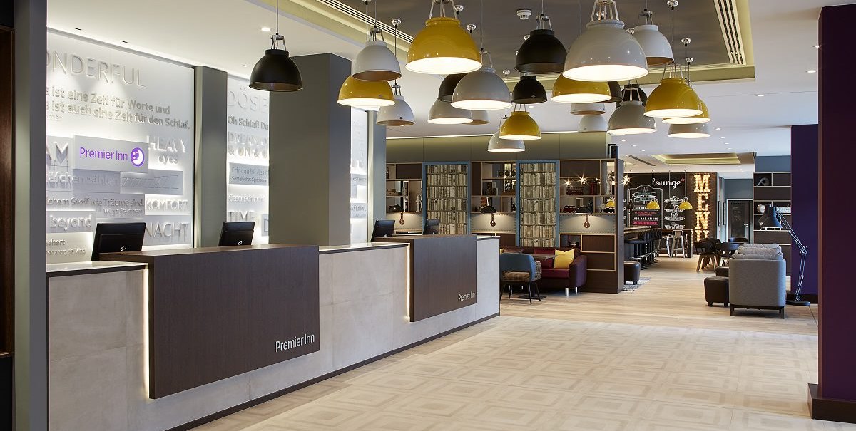 Moderne Hotel-Lobby mit Rezeption, stilvollen Hängelampen und gemütlichem Sitzbereich. Im Hintergrund ist ein beleuchtetes Menü-Schild zu sehen., © Premier Inn Moderne Hotel-Lobby mit Rezeption, stilvollen Hängelampen und gemütlichem Sitzbereich. Im Hintergrund ist ein beleuchtetes Menü-Schild zu sehen., © Premier Inn