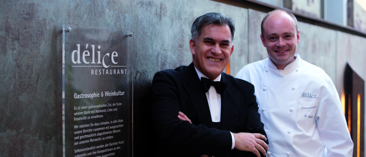 Zwei Männer stehen vor einem Schild des Délice Restaurants. Einer trägt einen Anzug, der andere eine Kochjacke., © Delice Zwei Männer stehen vor einem Schild des Délice Restaurants. Einer trägt einen Anzug, der andere eine Kochjacke., © Delice