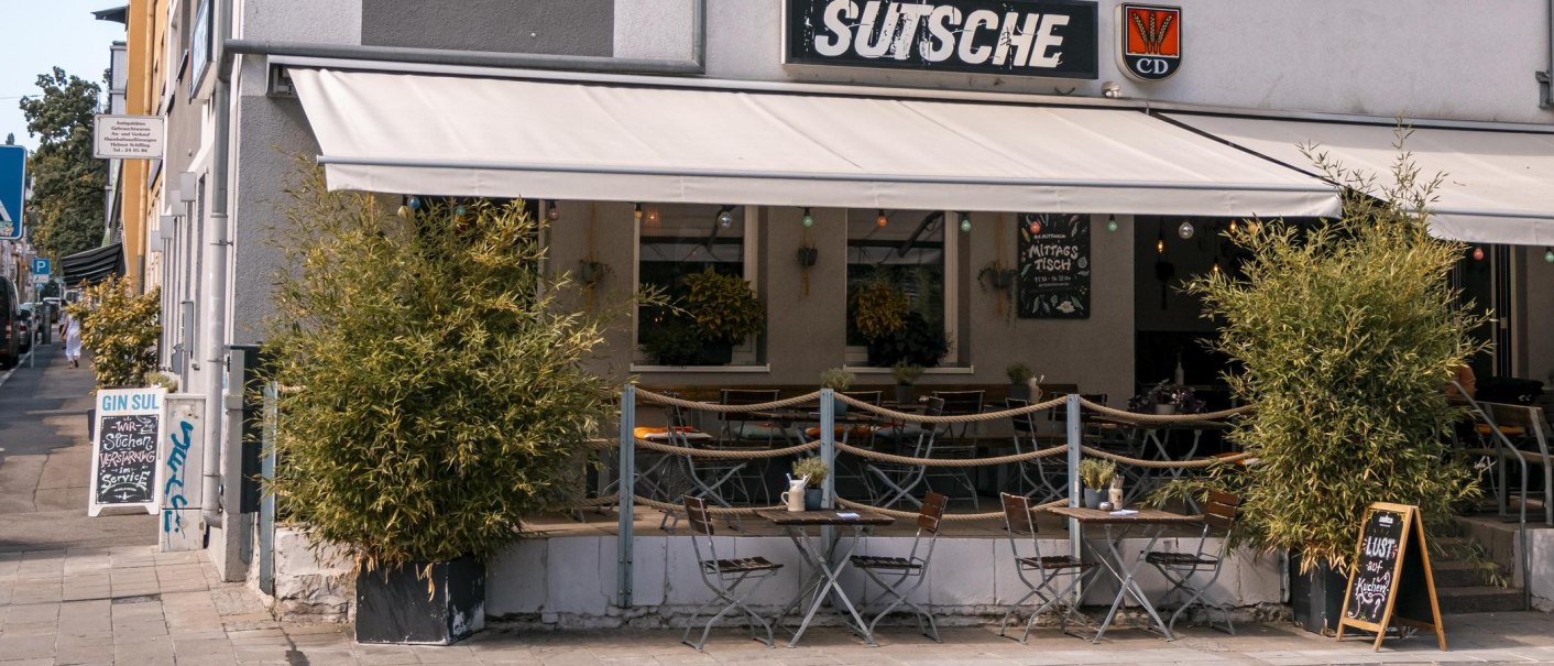Café 'Sutsche' mit Außenbereich, Tischen und Stühlen auf dem Gehweg, umgeben von Pflanzen., © SMG, Sarah Schmid Café 'Sutsche' mit Außenbereich, Tischen und Stühlen auf dem Gehweg, umgeben von Pflanzen., © SMG, Sarah Schmid