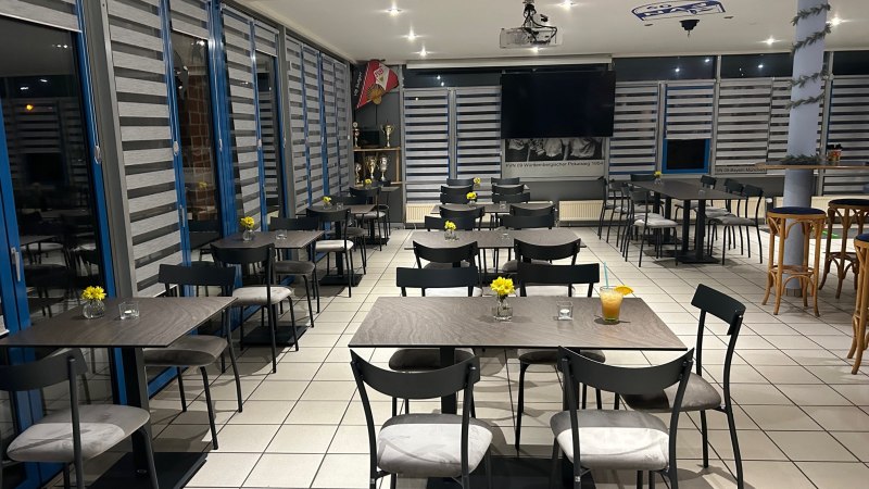 Ein modernes Bistro mit grauen Tischen und St&uuml;hlen, dekoriert mit gelben Blumen. Im Hintergrund sind Troph&auml;en und ein gro&szlig;er Bildschirm zu sehen., &copy; Bistro09