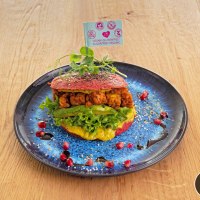 Ein veganer Burger mit Salat und Avocado auf einem blauen Teller, dekoriert mit Granatapfelkernen und Sesam. Ein F&auml;hnchen weist auf vegane, glutenfreie Zutaten hin., &copy; cEnergeticLife