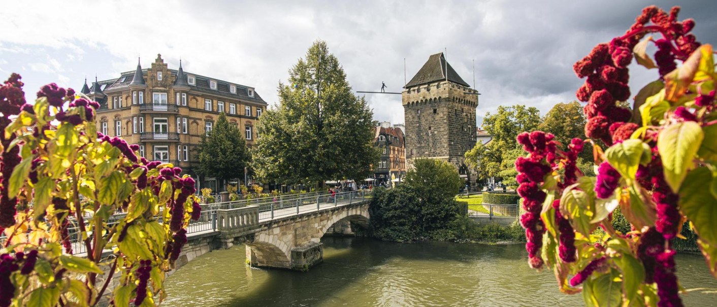 Der Schelztorturm in Esslingen, umgeben von historischen Gebäuden und einer Brücke über einen Fluss. Im Vordergrund sind bunte Blumen zu sehen., © SMG, Sarah Schmid Der Schelztorturm in Esslingen, umgeben von historischen Gebäuden und einer Brücke über einen Fluss. Im Vordergrund sind bunte Blumen zu sehen., © SMG, Sarah Schmid
