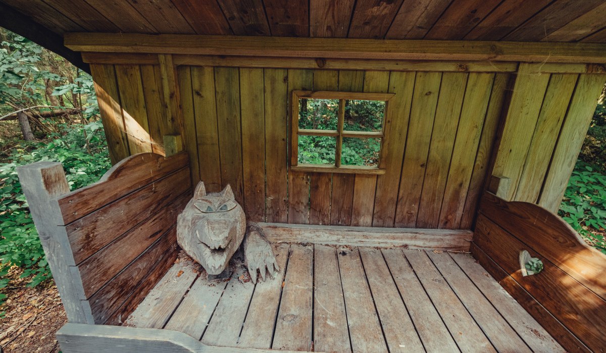 Eine Holzhütte im Märchenwald mit einer geschnitzten Wolfsskulptur auf einem Holzbett. Ein kleines Fenster zeigt den Wald im Hintergrund., © Tourismus Ostalb Eine Holzhütte im Märchenwald mit einer geschnitzten Wolfsskulptur auf einem Holzbett. Ein kleines Fenster zeigt den Wald im Hintergrund., © Tourismus Ostalb
