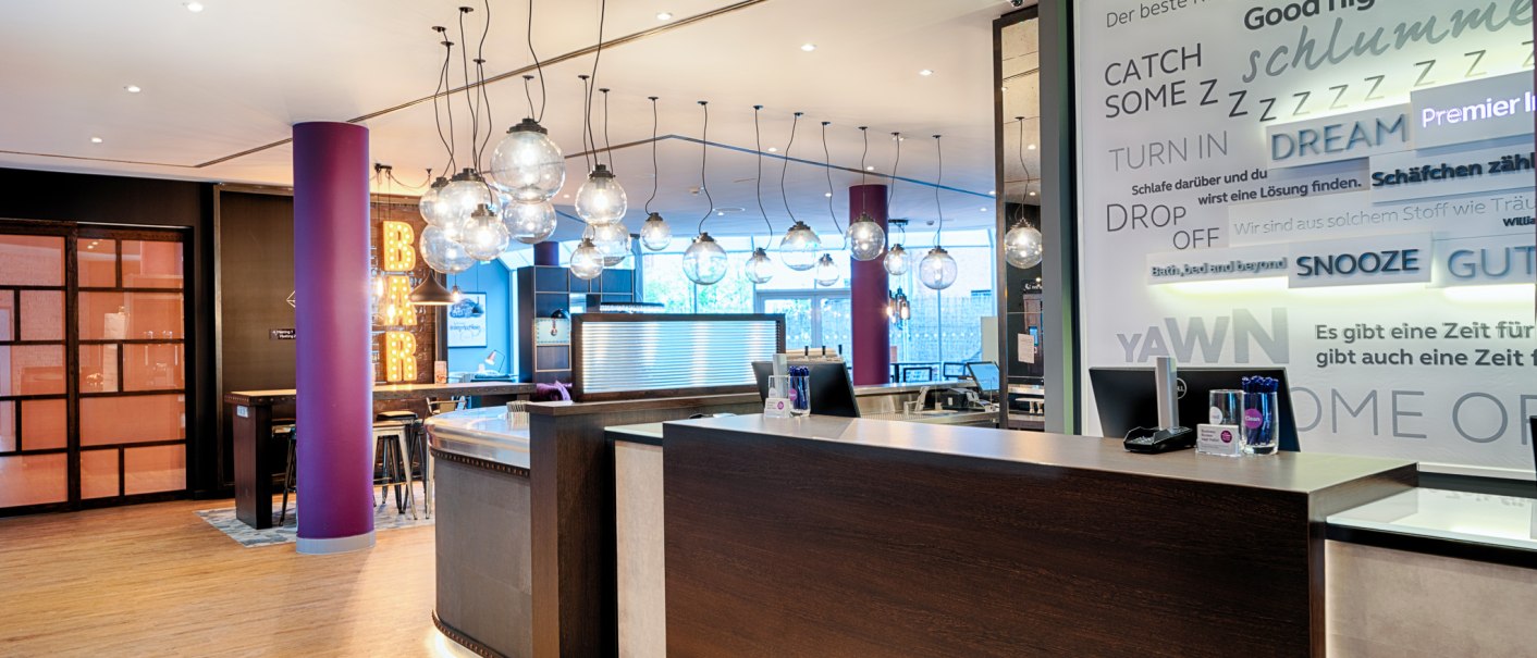Moderne Hotelrezeption mit hängenden Glaskugelleuchten. Im Hintergrund ist eine Bar mit leuchtendem Schriftzug zu sehen. Die Wände sind mit Texten dekoriert., © Premier Inn Germany