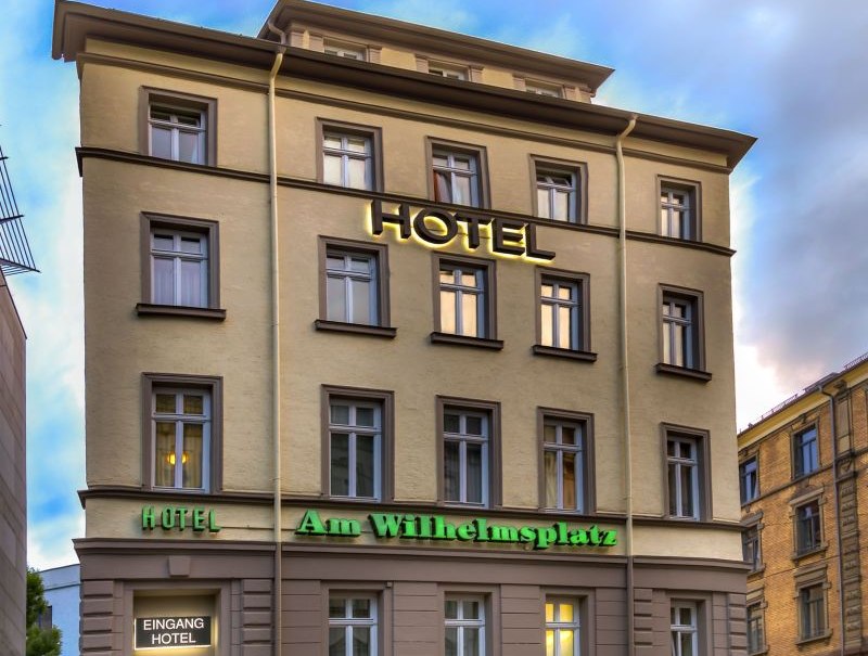 Mehrstöckiges Hotel mit der Aufschrift 'Hotel Am Wilhelmsplatz', davor ein geparktes Auto. Der Himmel ist bewölkt., © TOMAS