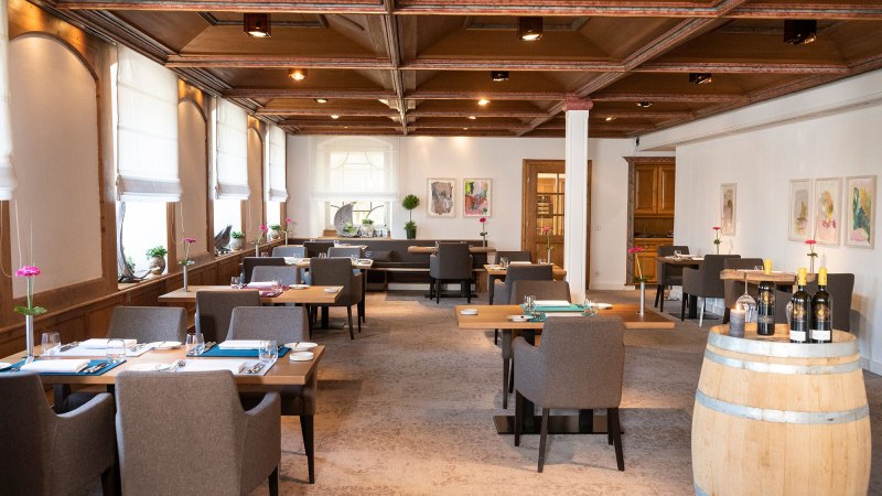 Elegantes Restaurant mit Holztischen, grauen Stühlen und einem Weinfass. Helle Fenster und moderne Kunst an den Wänden schaffen eine einladende Atmosphäre., © Schlosshotel Monrepos Elegantes Restaurant mit Holztischen, grauen Stühlen und einem Weinfass. Helle Fenster und moderne Kunst an den Wänden schaffen eine einladende Atmosphäre., © Schlosshotel Monrepos