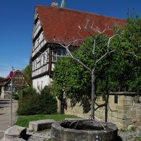 Ein Brunnen mit einer Baumskulptur aus Metall vor einem Fachwerkhaus. Der Himmel ist klar und blau, umgeben von gr&uuml;ner Vegetation., &copy; RadL Leonberg