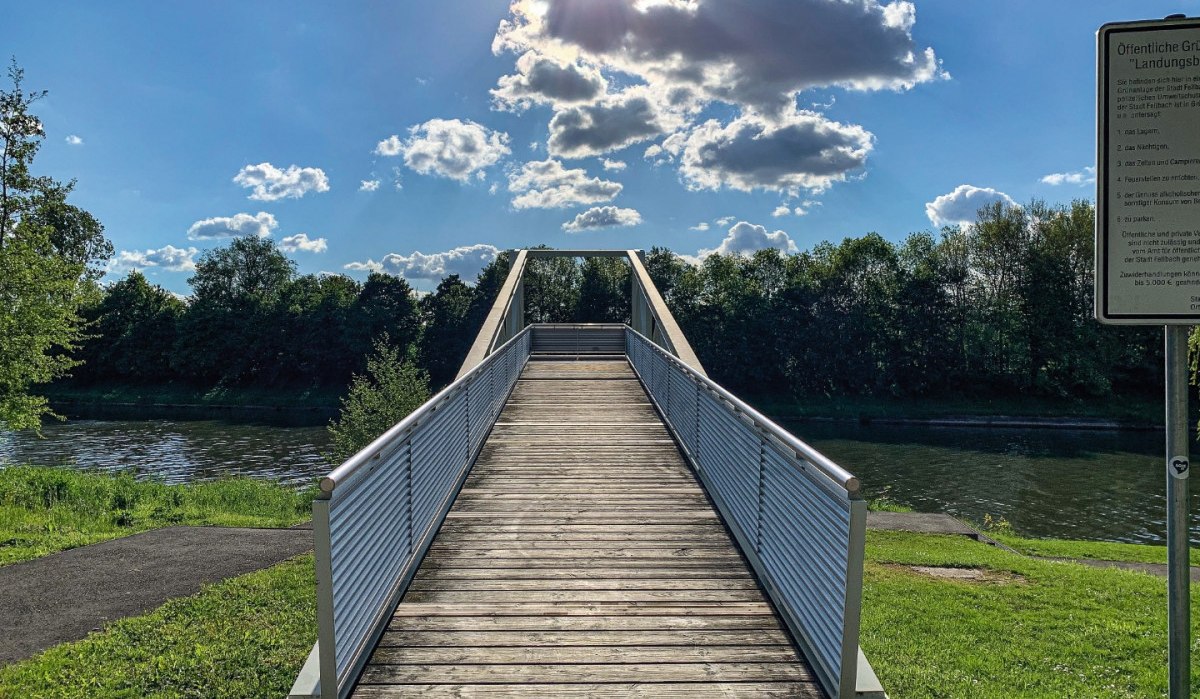Eine Holzbrücke führt über einen Fluss, umgeben von grüner Landschaft. Der Himmel ist blau mit einigen Wolken, die Sonne scheint hell., © Remstal Tourismus e.V.