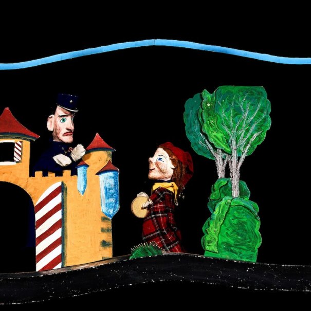 Zwei Puppenfiguren in einem Theaterstück. Eine Figur steht vor einem Schloss, die andere trägt einen Korb. Im Hintergrund sind Bäume zu sehen., © Prinzessin Gisela Theater