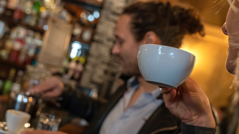 Zwei Personen in einem Caf&eacute;. Eine Person trinkt aus einer Tasse, w&auml;hrend die andere Kaffee zubereitet. Im Hintergrund sind Regale mit Flaschen zu sehen., &copy; Stuttgart Marketing  GmbH, Fotografin Martina Denker