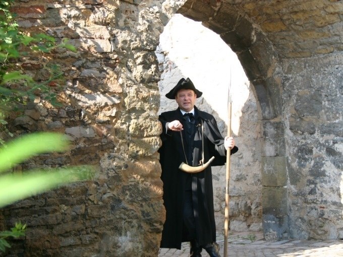 Ein Mann in historischer Kleidung mit Horn und Stab steht unter einem steinernen Torbogen und zeigt auf etwas., © WTM GmbH Waiblingen