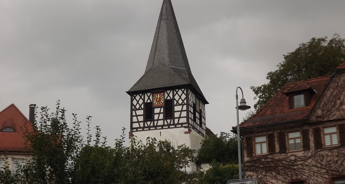 Ein Fachwerkturm mit Uhr und spitzem Dach erhebt sich vor einem bewölkten Himmel, umgeben von Bäumen und Häusern., © Natur.Nah. Schönbuch & Heckengäu Ein Fachwerkturm mit Uhr und spitzem Dach erhebt sich vor einem bewölkten Himmel, umgeben von Bäumen und Häusern., © Natur.Nah. Schönbuch & Heckengäu