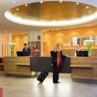 Eine moderne Hotelrezeption mit zwei Angestellten und einer Frau mit Koffer. Im Hintergrund sitzen Gäste auf Sofas. Die Einrichtung ist stilvoll und einladend., © Mercure Stuttgart Airport Messe