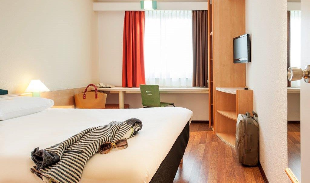 Ein gemütliches Hotelzimmer mit Doppelbett, Schreibtisch, Stuhl und Gepäck. Ein Fenster mit Vorhängen lässt Tageslicht herein., © TOMAS