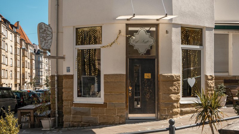Eingang des Café Seyffer’s an einer Straßenecke, dekoriert mit goldenen Girlanden und Pflanzen. Die Straße ist von Wohnhäusern gesäumt., © SMG, Sarah Schmid Eingang des Café Seyffer’s an einer Straßenecke, dekoriert mit goldenen Girlanden und Pflanzen. Die Straße ist von Wohnhäusern gesäumt., © SMG, Sarah Schmid
