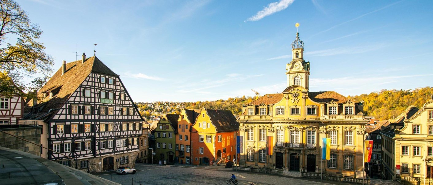 Der Marktplatz von Schwäbisch Hall zeigt Fachwerkhäuser und ein barockes Gebäude unter blauem Himmel. Die Herbstbäume im Hintergrund leuchten in warmen Farben., © Stuttgart-Marketing GmbH, Sarah Schmid Der Marktplatz von Schwäbisch Hall zeigt Fachwerkhäuser und ein barockes Gebäude unter blauem Himmel. Die Herbstbäume im Hintergrund leuchten in warmen Farben., © Stuttgart-Marketing GmbH, Sarah Schmid
