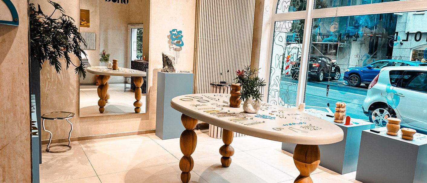 Modernes Studio mit Holztisch, Spiegel und Dekorationen. Große Fensterfront mit Blick auf die Straße. Pflanzen und Kunstobjekte als Dekoration., © Goldmarlen