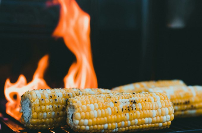 Drei Maiskolben liegen auf einem Grillrost, während im Hintergrund Flammen lodern. Perfekt für ein Veggie BBQ., © Cool-Tours StattReisen